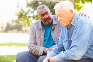 Two older men sit next to each other. One comforts the other.